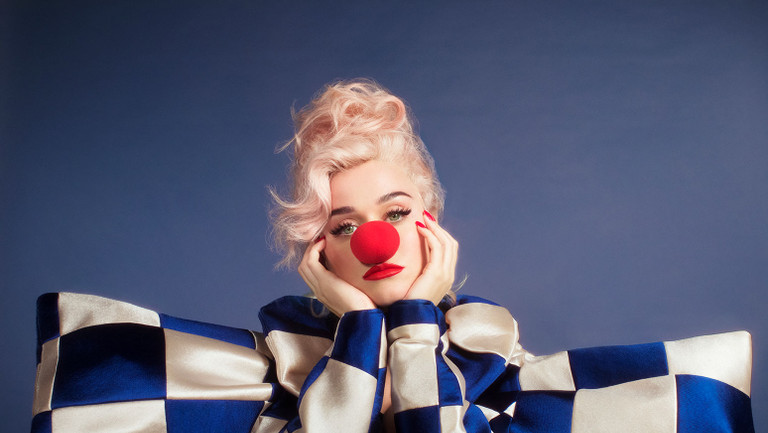 Foto de Katy Perry com roupa xadrez azul e branca larga, nariz de palhaço, sobre um fundo azul. Cantora prepara um documentário sobre maternidade real