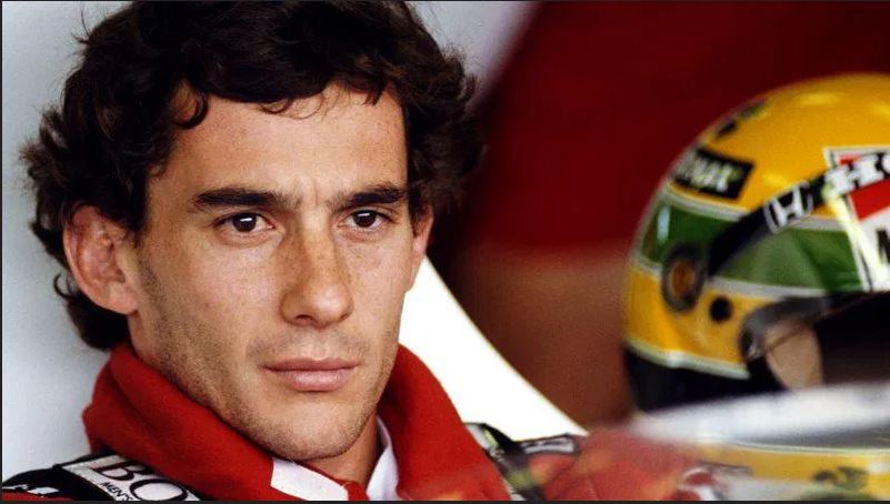 Piloto Ayrton Senna em close com olhar sério e concentrado com a roupa vermelha da Ferrari e seu capacete amarelo ao fundo