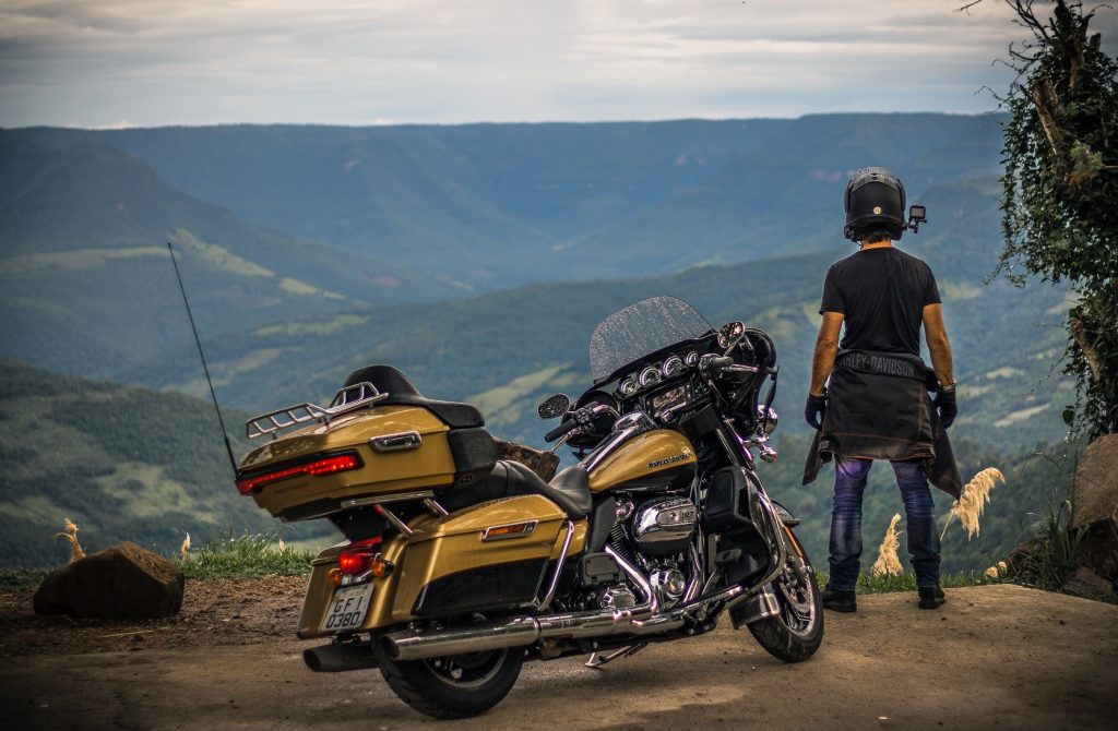 Viajar pelo Brasil: Motoqueiro de pé ao lado de uma Harley Davidson amarelada virado para uma paisagem montanhosa esverdeadas