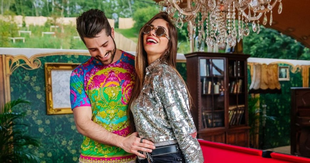 DJ Alok com blusa colorida acariciando a barriga de Romana Novaes, de blusa prateada e saia preta. O casal está sobre um fundo colorido e sorrindo