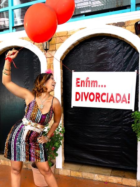 Ensaio de divorciada: Carol de vestido colorido segurando balões vermelhos e sorrindo ao lado de uma faixa escrito Enfim...Divorciada.