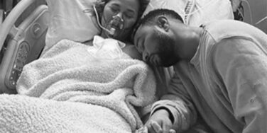 Chrissy Teigen deitada em uma cama de hospital desacordada com o marido John Legend sentado ao lado, segurando sua mão. Os dois parecem tristes. A foto é em preto e branco.