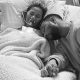 Chrissy Teigen deitada em uma cama de hospital desacordada com o marido John Legend sentado ao lado, segurando sua mão. Os dois parecem tristes. A foto é em preto e branco.