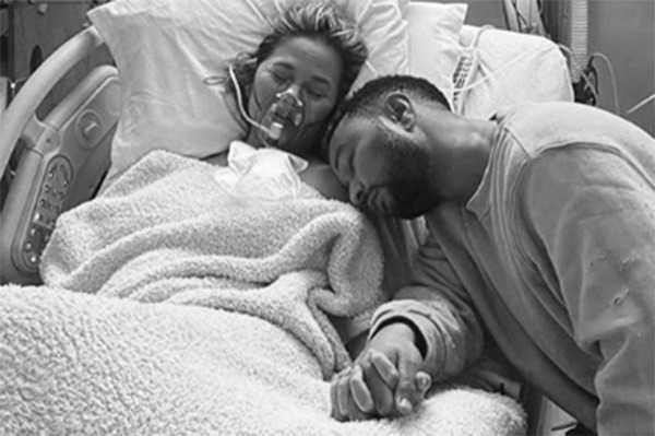 Chrissy Teigen deitada em uma cama de hospital desacordada com o marido John Legend sentado ao lado, segurando sua mão. Os dois parecem tristes. A foto é em preto e branco.