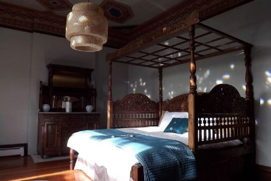 Quarto com uma cama no centro, com um dossel de madeira entalhada, um lustre no teto e um aparador ao lado.