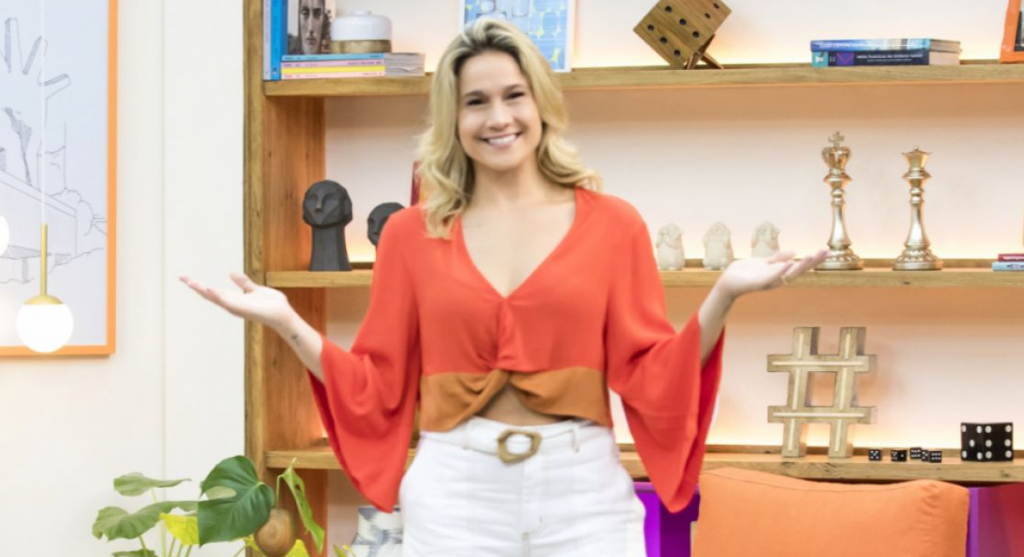 Luciano Huck no Domingão: Imagem de Fernanda Gentil de blusa laranja e calça branca, sorrindo com os braços abertos